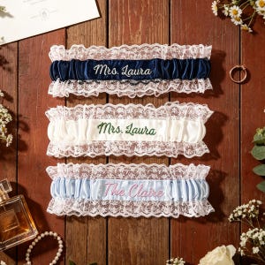 May include: Three elasticized lace garters featuring satin bands. The top garter is navy blue with white embroidery reading Mrs. Laura. The middle garter is white with green embroidery reading Mrs. Laura. The bottom garter is light blue with pink embroidery reading The Claire.