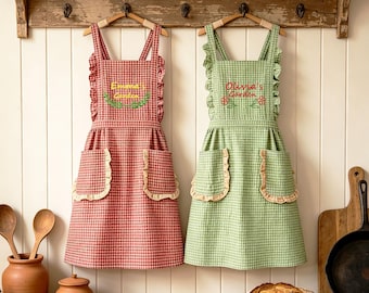 May include: Two aprons with ruffled straps and pockets, one in red gingham with 'Emma's Garden' embroidered in yellow, and one in green gingham with 'Olivia's Garden' embroidered in red. Both feature a checkered pattern and ruffled trim.