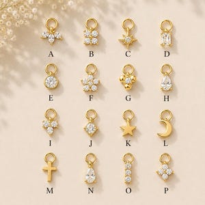 May include: A collection of sixteen gold-toned metal charms labelled A through P, featuring various shapes including stars, moons, crosses, flowers, and geometric designs, many accented with clear, sparkling cubic zirconia stones and jump rings.