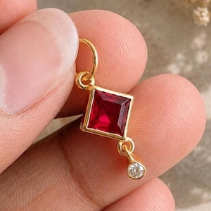 Pode incluir: Um pingente de ouro com uma pedra preciosa vermelha escura de corte quadrado montada em forma de diamante, com uma pequena pedra clara circular pendurada na ponta inferior. O pingente inclui um anel de ouro no topo para fixação.