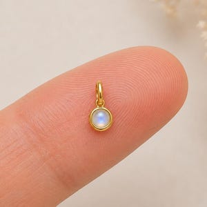 May include: A small, round gold pendant featuring a bezel-set, iridescent moonstone cabochon. The charm has a simple circular bail at the top for attachment to a chain, showcasing a polished metallic finish and a smooth, glowing gemstone surface.