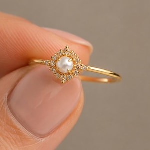 May include: A gold ring featuring a central round white pearl set within a square-shaped halo of small clear stones. The thin, polished gold band supports the decorative setting, which is centered on the ring's top surface for a balanced look.