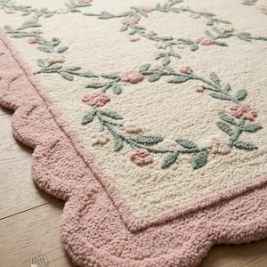 May include: A rectangular tufted rug with a cream-coloured centre featuring a repeating pattern of sage green vines and small pink flowers. The rug has a wide, scalloped border in a solid dusty pink colour. A small logo with the letters IRW is visible.