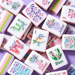 May include: A collection of white rectangular mahjong tiles featuring colorful engraved designs including floral arrangements, a bird on a branch, the letters W and U, the word Joker, the word Bam, and striped patterns in pink, green, and white.