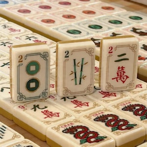 May include: A set of off-white mahjong tiles featuring engraved designs. Three tiles stand upright in the foreground: one with two green circles, one with green bamboo stalks, and one with the red Chinese character for two and the number 2.