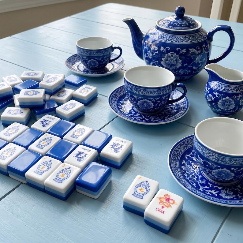 May include: A blue and white porcelain tea set including a teapot, three teacups with saucers, and a creamer, accompanied by a collection of rectangular mahjong tiles featuring blue and white floral patterns and the text Joker and Crak.