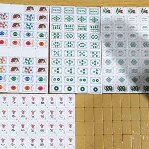 May include: A complete set of white mahjong tiles featuring various colorful illustrations including fish, flowers, green circular patterns, and red Chinese characters, alongside a group of plain tan-colored tiles arranged in a rectangular grid.