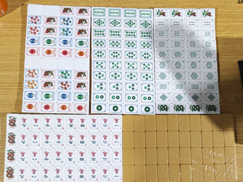 May include: A complete set of white mahjong tiles featuring various colorful illustrations including fish, flowers, green circular patterns, and red Chinese characters, alongside a group of plain tan-colored tiles arranged in a rectangular grid.