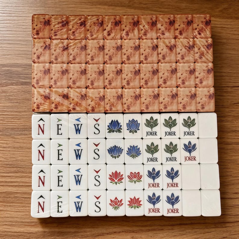 May include: A set of rectangular mahjong tiles arranged in rows. The top rows feature a brown marbled pattern. The bottom rows are white with printed designs including the letters N, E, W, S with directional arrows, lotus flowers, and Joker text.