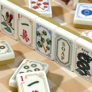 May include: A collection of white Mahjong tiles with gold-toned backs, featuring various colorful illustrations including a crane, red flowers, a green dragon, and the text SOAP and BAM, alongside tiles with Chinese characters and circular patterns.