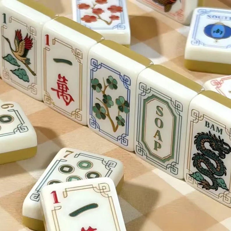 May include: A collection of white Mahjong tiles with gold-toned backs, featuring various colorful illustrations including a crane, red flowers, a green dragon, and the text SOAP and BAM, alongside tiles with Chinese characters and circular patterns.