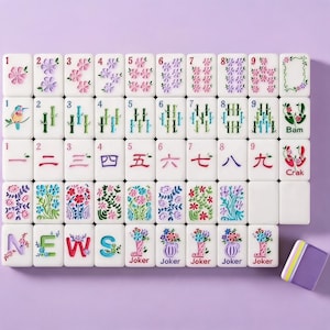 May include: A set of white mahjong tiles featuring colorful floral, bamboo, and character designs. The tiles display numbers one through nine, Chinese characters, directional letters spelling NEWS, and Joker tiles with floral boot illustrations.