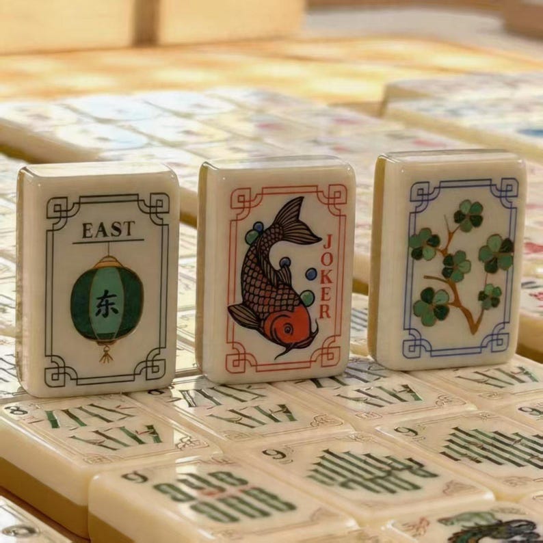 May include: Three off-white rectangular mahjong tiles featuring colorful illustrations. The left tile shows a green lantern with the text EAST, the center tile depicts a koi fish with the word JOKER, and the right tile displays a green floral branch.
