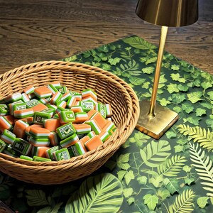 May include: A round woven wicker basket filled with numerous small rectangular mahjong tiles featuring orange, white, and green layers with green character engravings. A gold-toned metal lamp stands next to the basket on a green leaf-patterned surface.