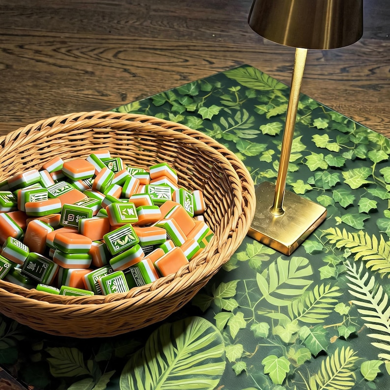 May include: A round woven wicker basket filled with numerous small rectangular mahjong tiles featuring orange, white, and green layers with green character engravings. A gold-toned metal lamp stands next to the basket on a green leaf-patterned surface.