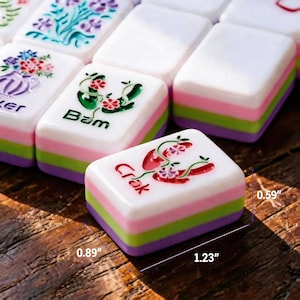 May include: A set of rectangular Mahjong tiles with a white top surface featuring colorful floral and character designs, including the text Bam and Crak. Each tile has layered pink, green, and purple sides, measuring 1.23 inches by 0.89 inches by 0.59 inches.