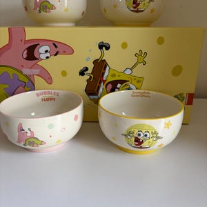 May include: A set of four ceramic bowls and a yellow box featuring SpongeBob SquarePants and Patrick Star characters. Two bowls display Patrick with colourful dots, while two display SpongeBob with yellow stars. Text includes BUBBLES HAPPY and SpongeBob SquarePants.