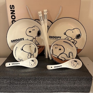May include: A Snoopy-themed dinnerware set including two round plates, two bowls, two spoons, and two pairs of chopsticks. Each piece is cream-coloured with black line art of the character Snoopy and the text SNOOPY and 2024 Peanuts Worldwide LLC.