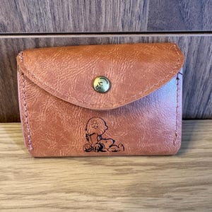 May include: A tan faux leather wallet with a curved flap closure secured by a brass-toned snap button. The front features a black line art illustration of a character reading a book while lying down next to a small dog.