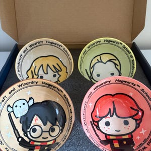 May include: A set of four round ceramic bowls featuring cartoon-style illustrations of characters from Hogwarts School of Witchcraft and Wizardry. Each bowl displays a different character with a unique background colour, including tan, light green, and pink.