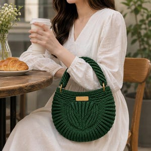 May include: A dark green, semi-circular crochet handbag featuring a thick, braided handle attached with gold-toned metal rings. The bag has a textured, ribbed pattern and a rectangular gold-toned metal plaque centered on the front exterior.