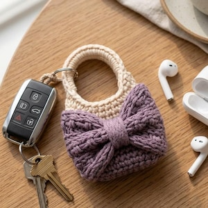 Pode incluir: Uma pequena bolsa de crochê com uma alça circular na cor creme e um laço texturizado em tom roxo empoeirado na frente. A bolsa está presa a um chaveiro de metal contendo uma chave de carro preta e duas chaves de metal prateado.