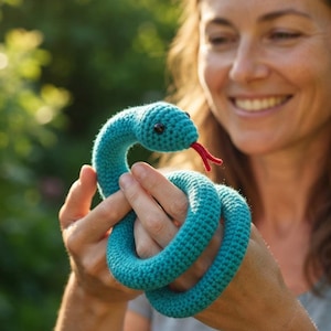 Puede incluir: Un juguete de serpiente tejido a ganchillo en color verde azulado con una superficie de hilo texturizado, cabeza redondeada con pequeños ojos de cuentas negras y una lengua bífida de hilo rojo que sobresale de la boca, enrollada en círculo.