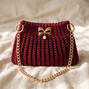 Puede incluir: Un bolso de mano de ganchillo en color burdeos intenso con un patrón de canalé texturizado. El bolso está adornado con un lazo de metal dorado centrado en la parte superior, que incluye una pequeña perla blanca. Tiene una correa de cadena dorada.