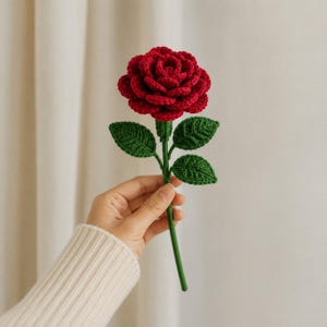 May include: A handcrafted crochet rose featuring a deep red, multi-layered flower head with a textured surface. The stem is a vibrant green, complemented by three detailed green leaves attached to the stalk, showcasing intricate yarn stitching.