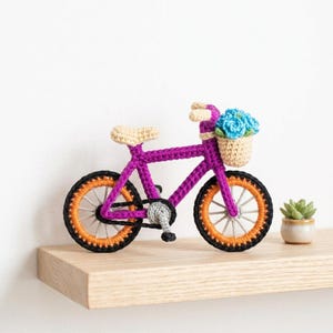May include: A crocheted bicycle figurine featuring a vibrant purple frame, orange wheels with black tires, and a beige seat. A small beige basket is attached to the handlebars, filled with bright blue crocheted flowers and green leaves.