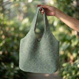 May include: A sage green crochet tote bag featuring a dense, textured stitch pattern with two integrated shoulder straps. The bag has a structured, open-top design with a consistent, uniform weave throughout the entire body and handle construction.