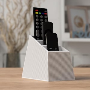 May include: A white, wedge-shaped remote control holder with a vertical ribbed texture on the exterior. The organiser features three tiered compartments of varying heights, currently holding one large remote and two smaller streaming device remotes.