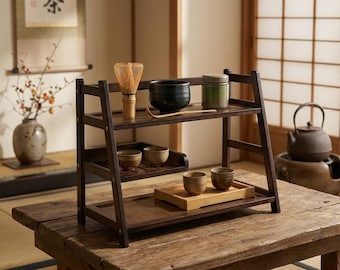 May include: A dark brown wooden tiered display shelf holding a bamboo matcha whisk, a black ceramic bowl, a green tea canister, and four small ceramic tea cups arranged on two levels, with one pair of cups resting on a light bamboo tray.