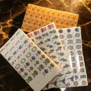 May include: A set of white mahjong tiles featuring colorful engraved designs including directional letters, floral patterns, bamboo stalks, circular symbols, and joker tiles with text, arranged in overlapping stacks on a dark surface.