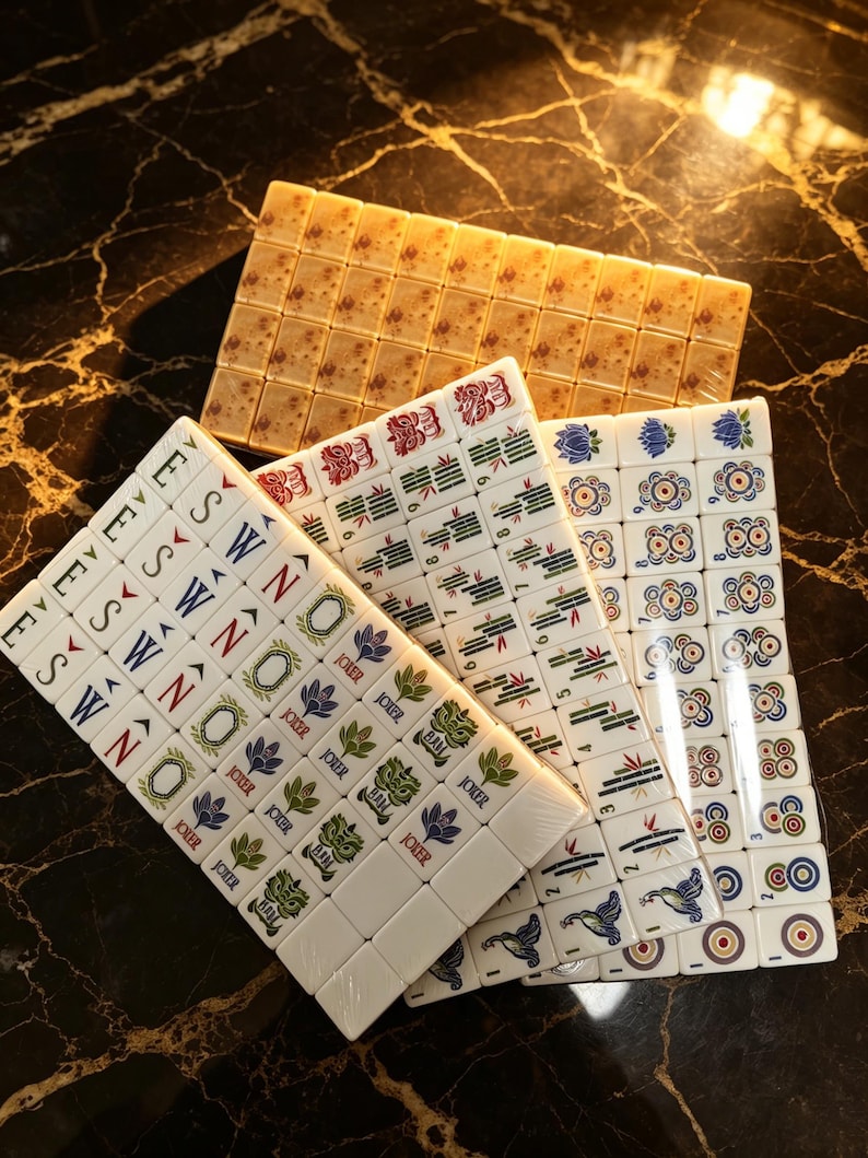 May include: A set of white mahjong tiles featuring colorful engraved designs including directional letters, floral patterns, bamboo stalks, circular symbols, and joker tiles with text, arranged in overlapping stacks on a dark surface.