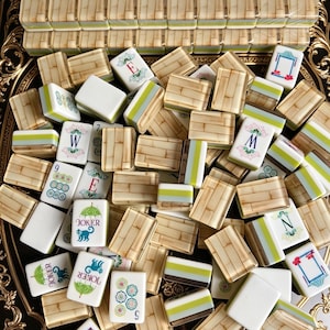 Puede incluir: Un juego de fichas de mahjong con reverso de patr&oacute;n de bamb&uacute; y cara blanca con detalles en verde y azul. Las fichas muestran varios s&iacute;mbolos, incluyendo las letras E, W, M, N, los n&uacute;meros 5, 6, 9 y un gr&aacute;fico de joker con un mono y paraguas.
