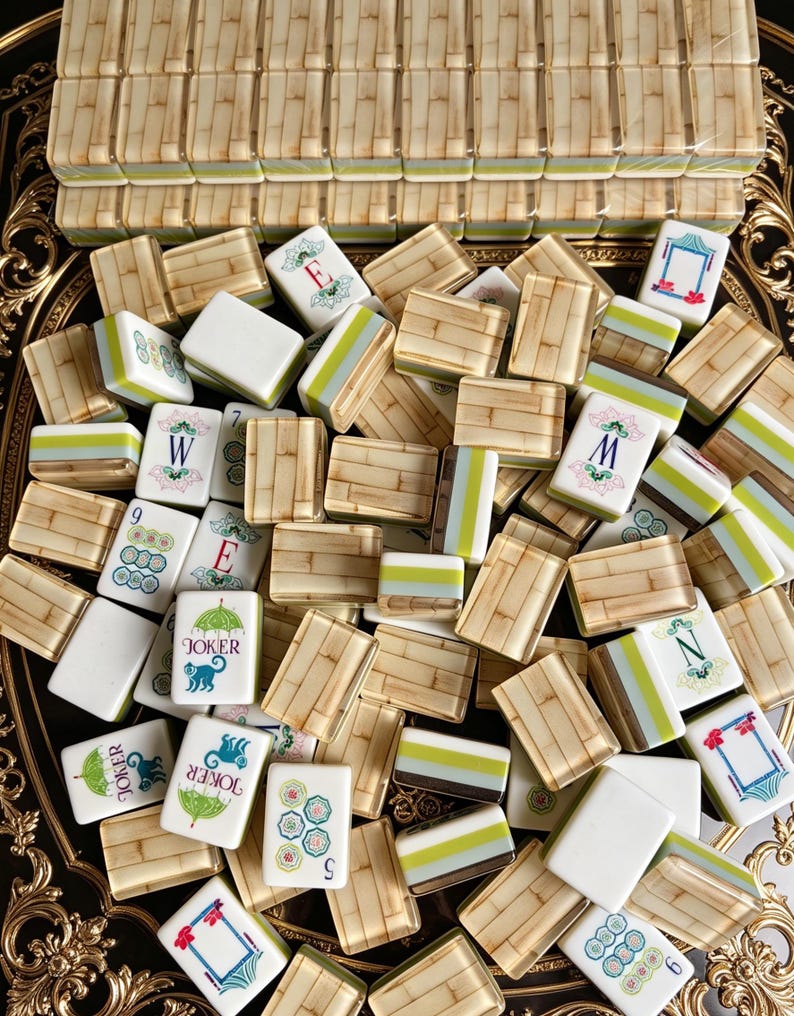 Puede incluir: Un juego de fichas de mahjong con reverso de patr&oacute;n de bamb&uacute; y cara blanca con detalles en verde y azul. Las fichas muestran varios s&iacute;mbolos, incluyendo las letras E, W, M, N, los n&uacute;meros 5, 6, 9 y un gr&aacute;fico de joker con un mono y paraguas.