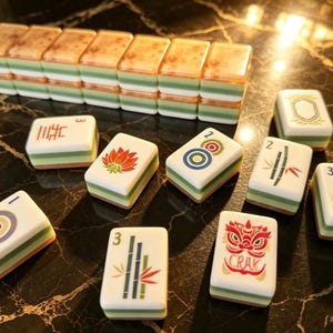 May include: A set of rectangular mahjong tiles featuring white faces with colorful illustrations including a red lotus, a green bamboo stalk, a red lion mask with the text CRAK, and various circular patterns, stacked with green and white layered sides.