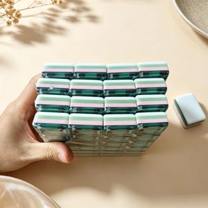 May include: A stack of rectangular mahjong tiles featuring a layered design with a dark green base, a thin pink middle stripe, and a light green top section. The tiles are arranged in a grid pattern, and one single matching tile sits separately nearby.