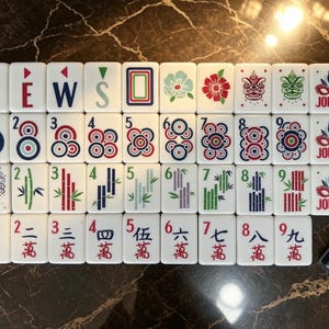 May include: A set of white Mahjong tiles arranged in four rows, featuring colorful designs including the letters N, E, W, S, floral patterns, circular motifs, bamboo stalks, Joker masks, and Chinese characters with red and blue ink accents.