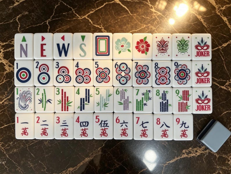 May include: A set of white Mahjong tiles arranged in four rows, featuring colorful designs including the letters N, E, W, S, floral patterns, circular motifs, bamboo stalks, Joker masks, and Chinese characters with red and blue ink accents.