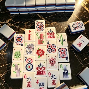 May include: A set of white Mahjong tiles featuring colorful graphic designs including floral patterns, bamboo stalks, masks, and Chinese characters. Some tiles are arranged in a grid, while others are stacked or placed individually.