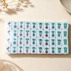 May include: A set of white rectangular mahjong tiles arranged in a grid, wrapped in clear plastic. Each tile features a colorful illustration of a plant in a pot, a bird, or a dragon, with numbers one through nine printed in the top left corner.