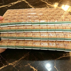 May include: A stack of four rectangular mahjong tile sets wrapped in clear plastic. Each tile features a mottled tan and brown top surface, a solid green middle layer, and a white base, arranged in neat rows within the protective packaging.