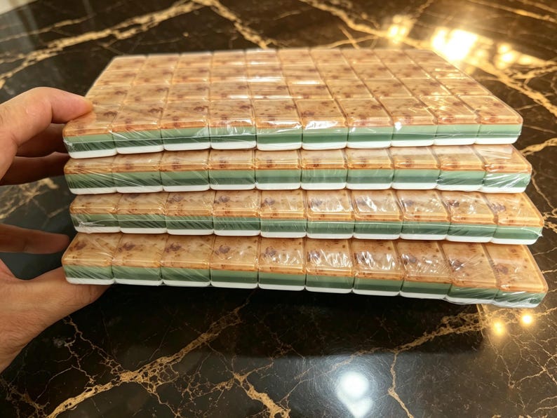 May include: A stack of four rectangular mahjong tile sets wrapped in clear plastic. Each tile features a mottled tan and brown top surface, a solid green middle layer, and a white base, arranged in neat rows within the protective packaging.