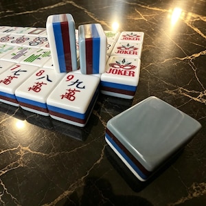 May include: A set of rectangular Mahjong tiles featuring white faces with red and green Chinese characters, blue and maroon striped sides, and Joker tiles with a red mask graphic. One tile is placed face down showing a solid grey back.