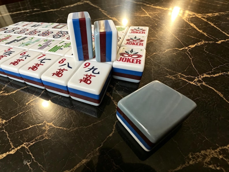 May include: A set of rectangular Mahjong tiles featuring white faces with red and green Chinese characters, blue and maroon striped sides, and Joker tiles with a red mask graphic. One tile is placed face down showing a solid grey back.