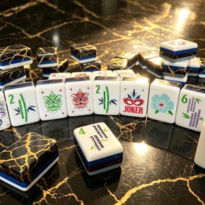 May include: A collection of Mahjong tiles featuring white faces with colorful illustrations of bamboo, flowers, dragons, and the word JOKER. The tiles have a dark blue base with gold veining patterns on the sides, arranged in a scattered display.