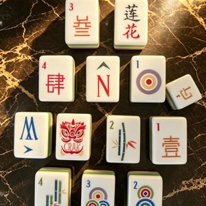 May include: A collection of twelve white Mahjong tiles with green backs, featuring various red, blue, and green engraved symbols, Chinese characters, numbers, and a red lion graphic with the text CRAK, arranged in a triangular formation.