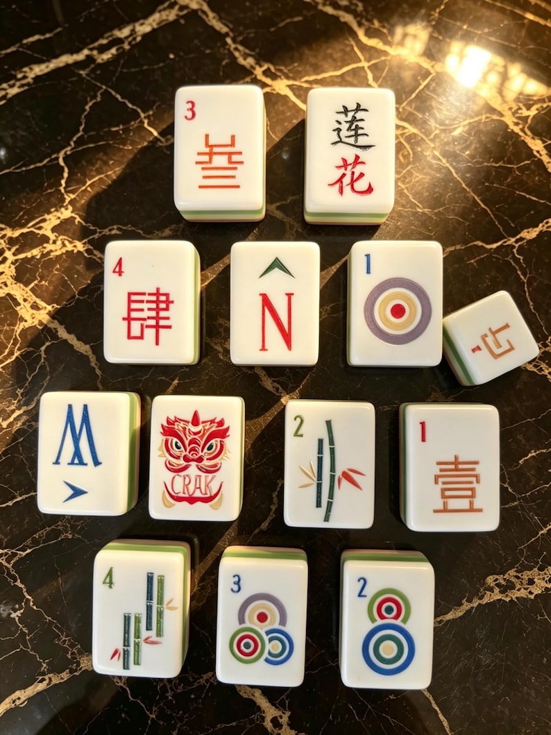 May include: A collection of twelve white Mahjong tiles with green backs, featuring various red, blue, and green engraved symbols, Chinese characters, numbers, and a red lion graphic with the text CRAK, arranged in a triangular formation.
